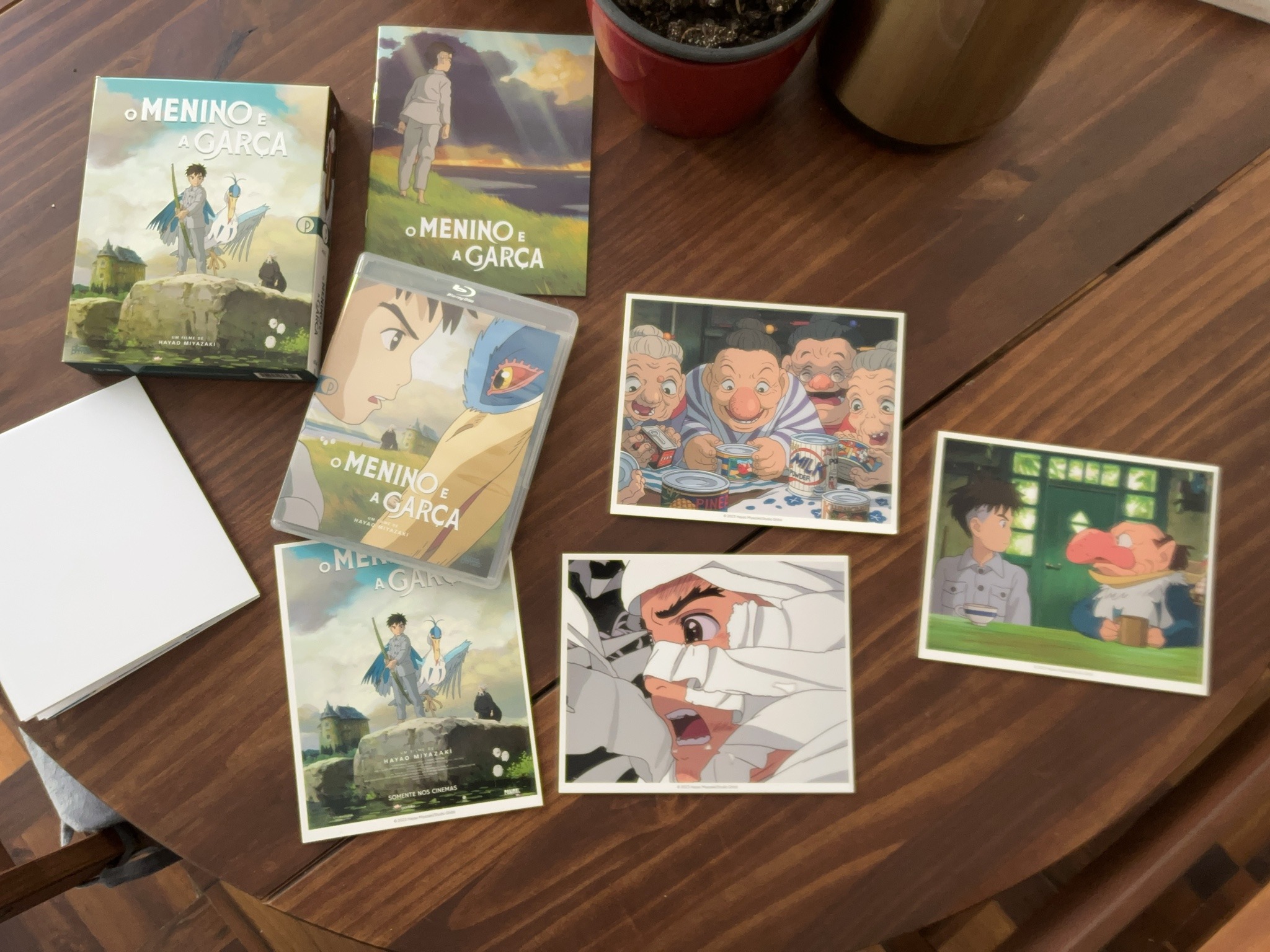 Os vários itens que fazem parte da edição de "O Menino e a Garça" em blu-ray estão espalhados em cima da mesa: vários postais com cenas, um caderno com um ensaio e o filme em si