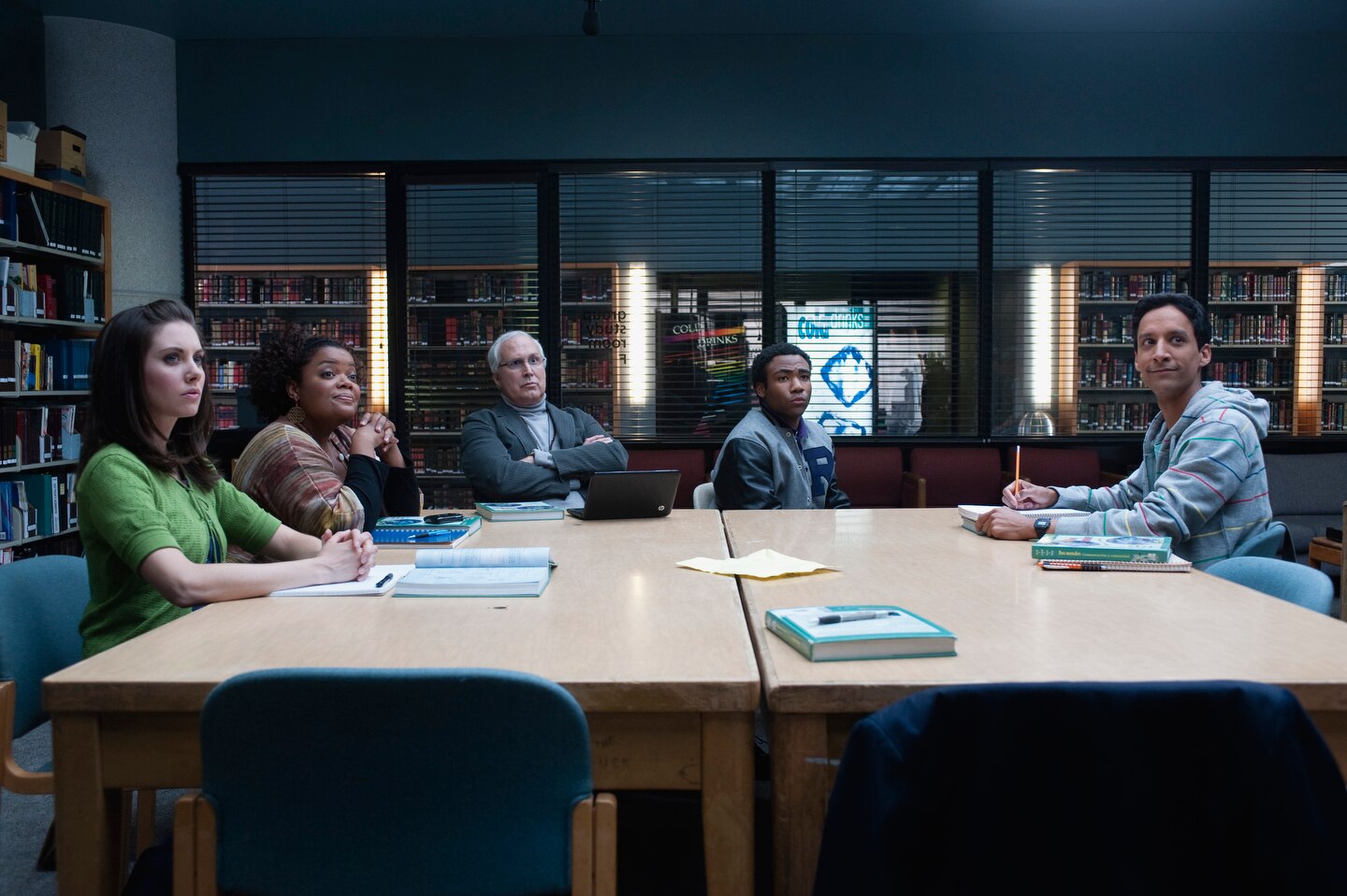 Cena de Community: o grupo de estudos está sentado ao redor da mesa na biblioteca olhando para Jeff, que está em pé (fora de cena)
