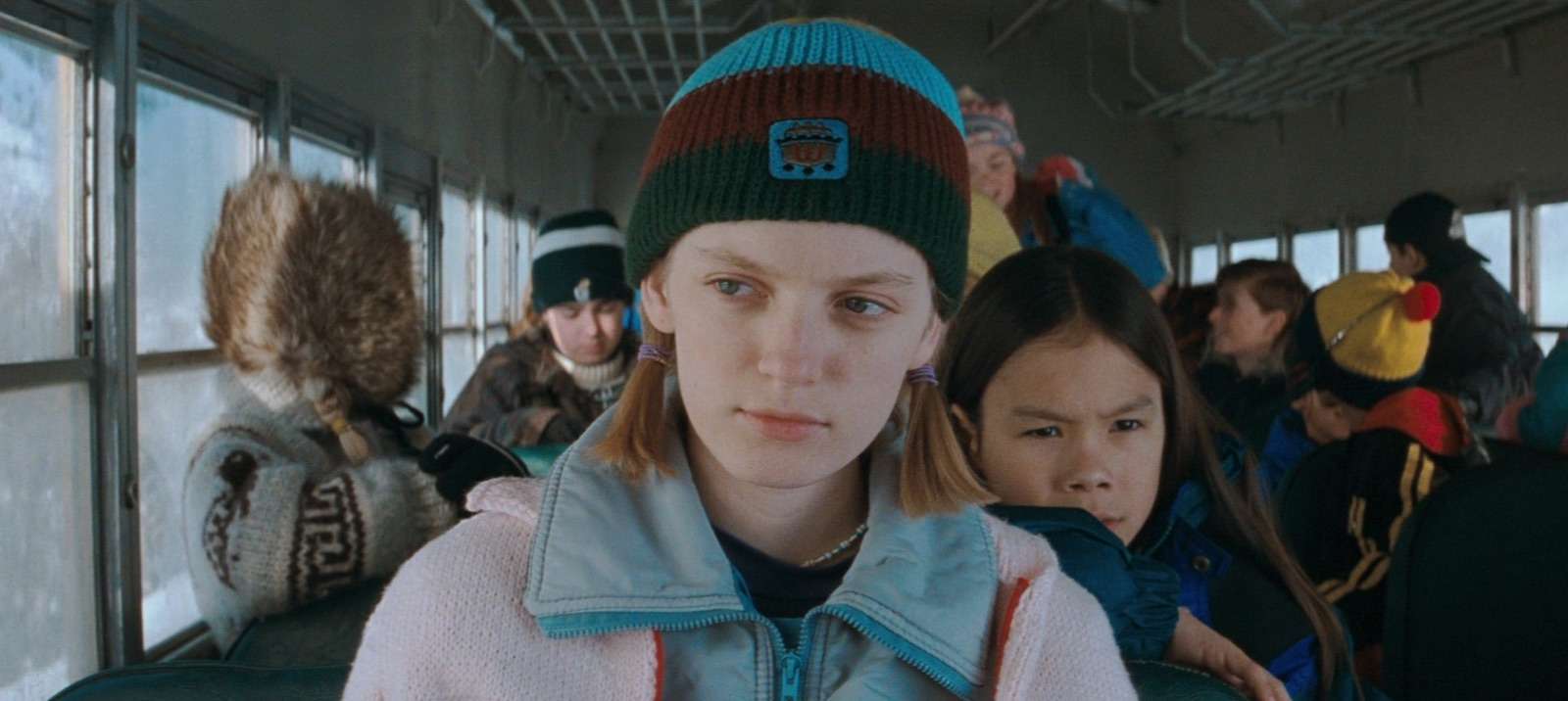 Crianças sentadas no ônibus escolar, em destaque está uma menina loira, com uma touca e roupa de frio