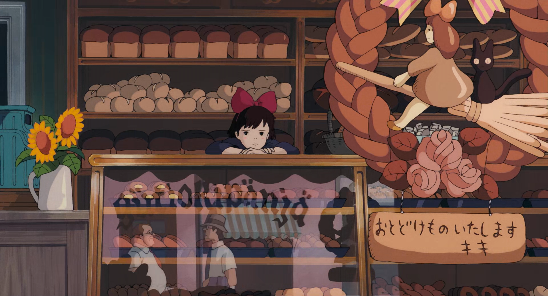 Kiki está apoiada no balcão da padaria olhando pela porta de vidro da loja, uma girlanda com uma bruxinha sentada numa vassoura com seu gato está pendurada na porta