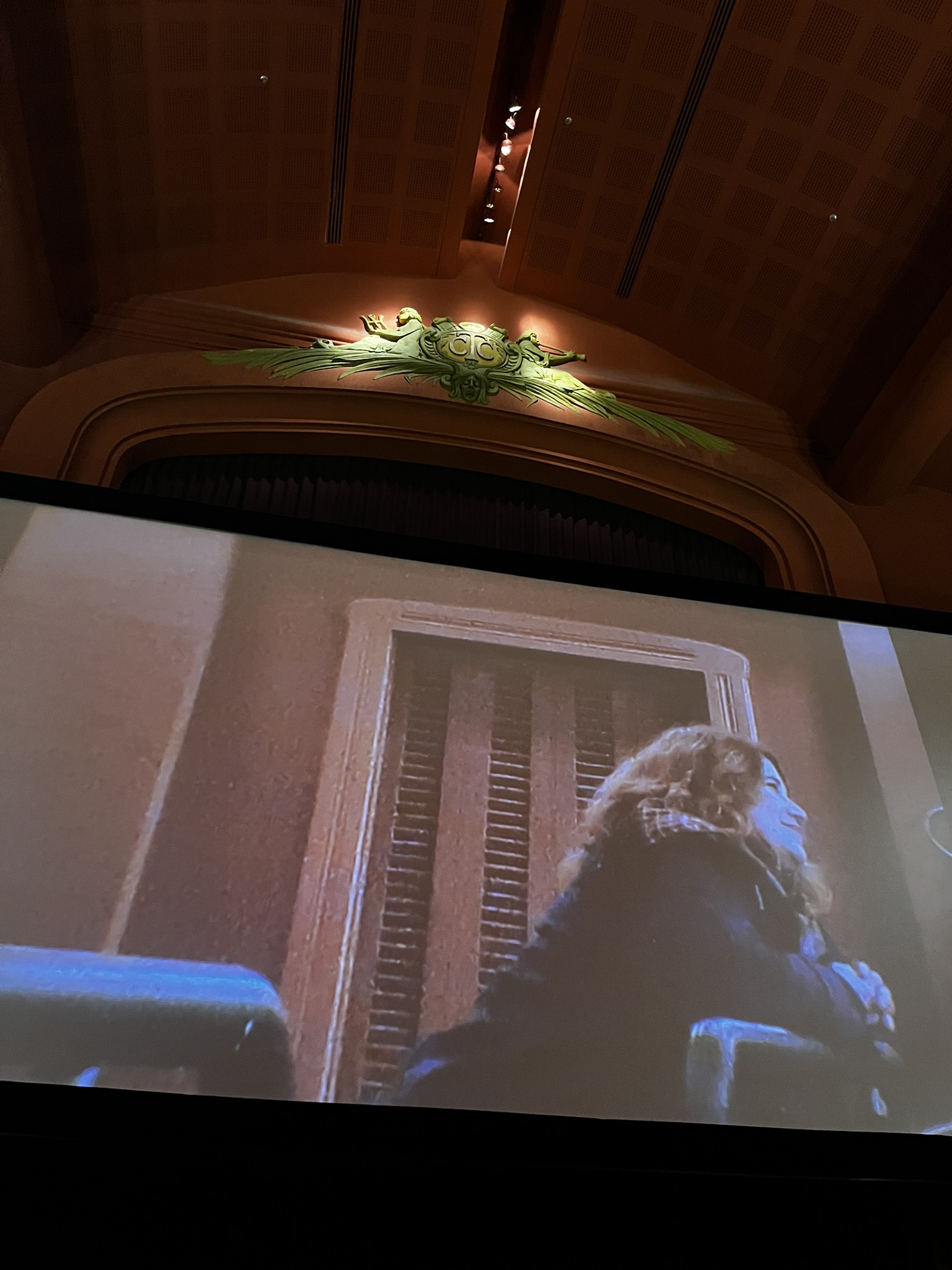 O telão da Cinemateca Capitólio, com uma foto da Tai na própria cinemateca, sorrindo debruçada no encosto de uma das poltronas, ouvindo alguém em alguma sessão de debates